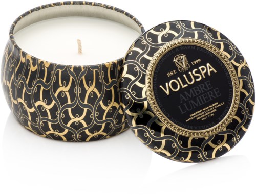A round candle in a decorative black and gold container with intricate patterns, lid is off showing the white wax inside.