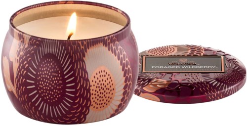 Een brandende geurkaars in een decoratieve roze en paarse glazen container. De deksel ligt ernaast.