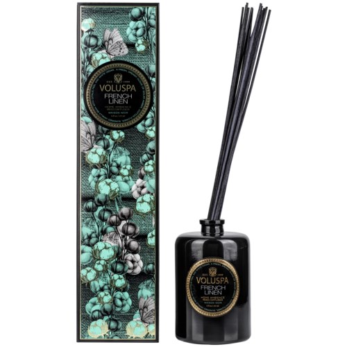 En svart diffuserflaska med svarta pinnar, bredvid en dekorerad box med botanisk design. Den är märkt 'Voluspa French Linen.'