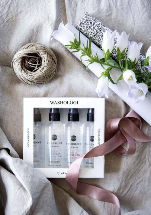 A gift set with four small bottles, surrounded by flowers, string, and a satin ribbon, on a soft cloth background.