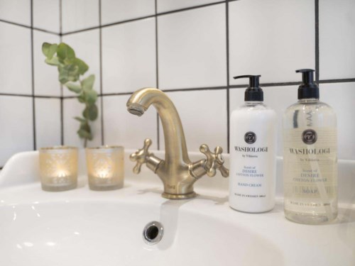 Ett handfat med två ljuslyktor, en växt och två flaskor med tvål eller lotion på en kaklad vägg.