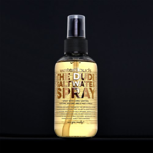 Een sprayfles met label "The Dude Salt Water Spray" voor haar, met een zwarte dop en doorzichtige body die geel licht laat zien.