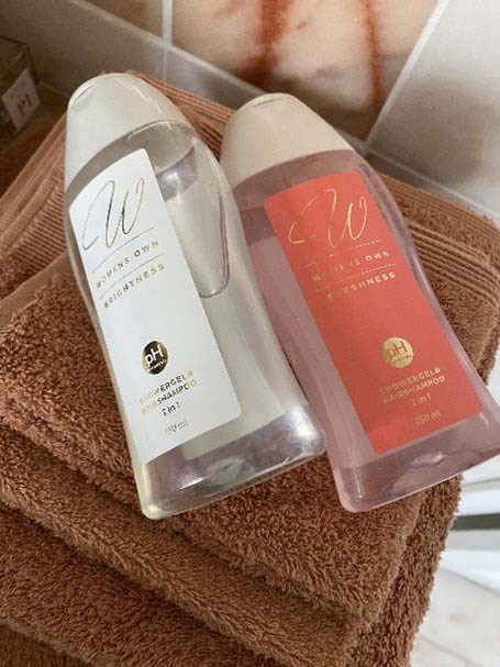 Zwei Shampooflaschen, eine weiße und eine rosa, liegen auf einem braunen Handtuch. Sie haben goldene und schwarze Etiketten.