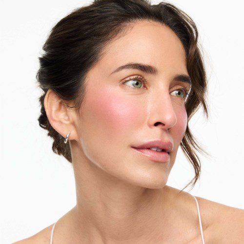 A woman with light skin and dark brown hair styled back, wearing minimal makeup and small hoop earrings.