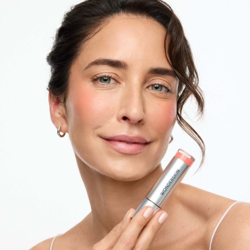 A woman with smooth skin holding a skincare product near her face, smiling subtly with a plain white background.