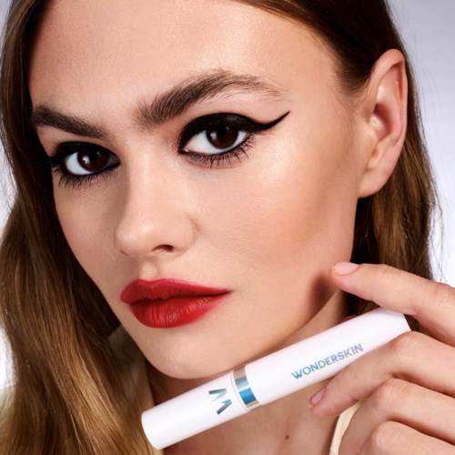 A woman with bold makeup, holding a skin care product labeled 'Wonderskin' near her face.