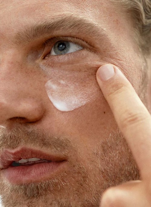 Een man brengt crème aan onder zijn oog en raakt het gebied aan met zijn vinger. Close-up van huidverzorgingsroutine.