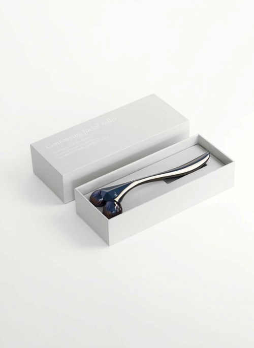 En ansiktsrullare med ett metallhandtag och två stenar inne i en vit box. Boxen är öppen. Rullaren är silver och mörkblå.