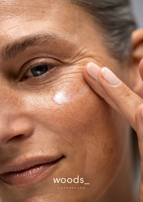 A woman applies cream under her eye, with visible wrinkles and clear skin. There is a logo 'woods COPENHAGEN' at the bottom.
