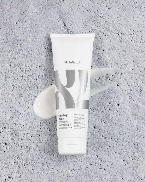 En hvit tube med Grazette bonding mask ligger på en teksturert grå betongoverflate med litt av kremen spredt i nærheten.