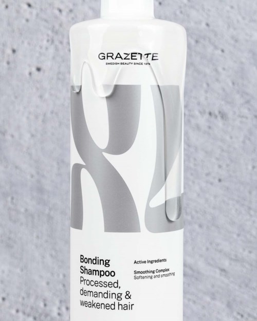 En vit flaska med Grazeite Bonding Shampoo med grå abstrakt design på etiketten, placerad mot en grå texturerad bakgrund.