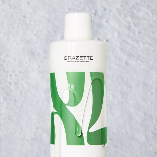 Eine weiße Flasche mit grünen Formen und schwarzem Text, der "GRAZETTE" sagt, auf grauem Hintergrund.