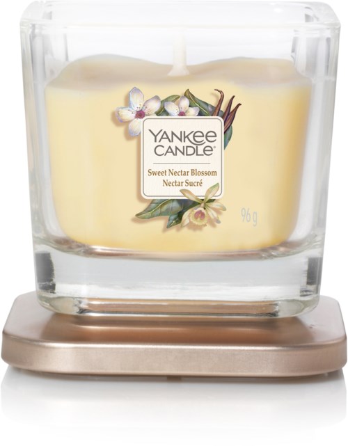 En Yankee Candle-kruka med ett gult ljus inuti, dekorerad med en blomma och vaniljbönor, på en metallbas.