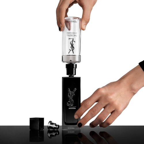 En hand håller ett klart glas med YSL-logotyp, och en annan hand trycker på en svart enhet.