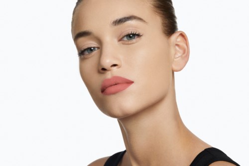 Een vrouw met een heldere huid, rode lipstick en een neutrale uitdrukking. Ze heeft bruin haar in een staart en draagt een zwart shirt.
