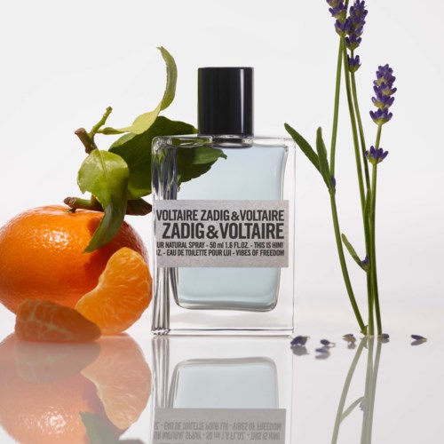 Een parfumfles omgeven door een sinaasappel, een groene vrucht en lavendelbloemen.