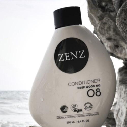 En vit flaska märkt ZENZ balsam med svart lock är placerad utomhus nära stenar och havet.