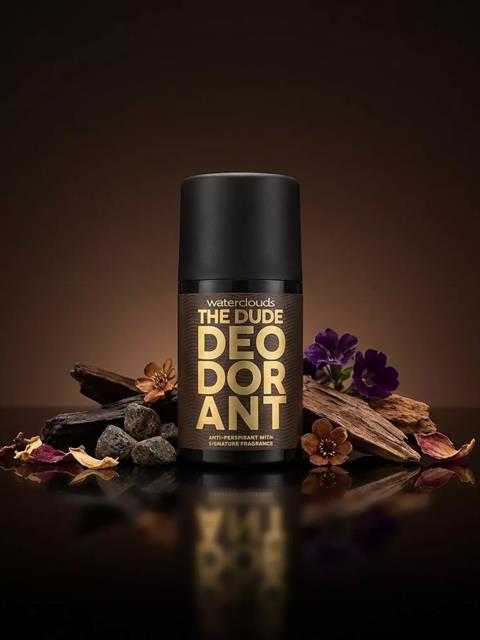 Billedet viser en sort deodorantdåse med guldtekst, omgivet af sten, træstykker og lilla blomster mod en mørk baggrund.