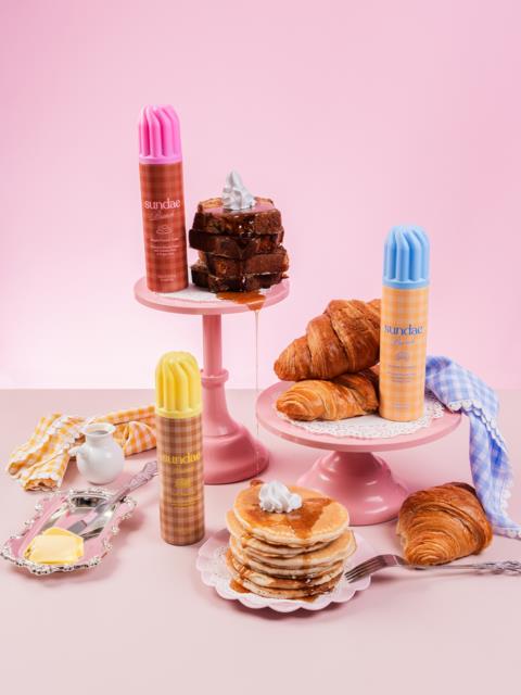 Ljust rosa bakgrund med staplade bakverk, croissanter, grädde och färgglada flaskor på rosa ställningar.