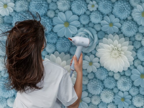 En kvinne tørker håret med en hvit hårføner foran en bakgrunn av blå og hvite blomster.
