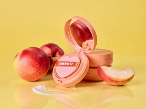 Een make-up case met spiegel, omringd door appels en perzikgekleurde productverpakkingen op een gele ondergrond.