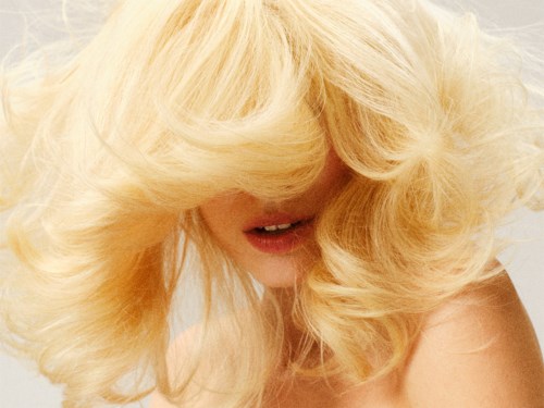 Een vrouw met lang, golvend blond haar bedekt een deel van haar gezicht, toont haar neus en half geopende lippen.