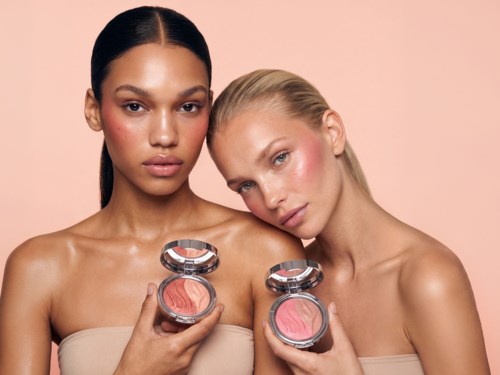 Två kvinnor håller öppna blushprodukter framför sig, visar sitt smink och skinande hud mot en rosa bakgrund.