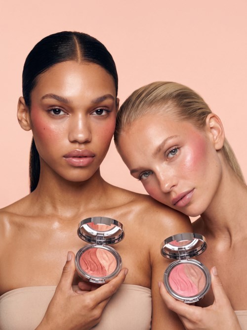 To kvinder holder i pink blush produkter, den ene med mørkt hår og den anden med blond, mod en lys pink baggrund.