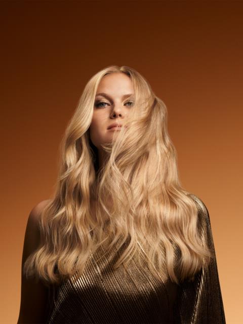 A woman with long, wavy blonde hair wearing a gold top against a brown background.