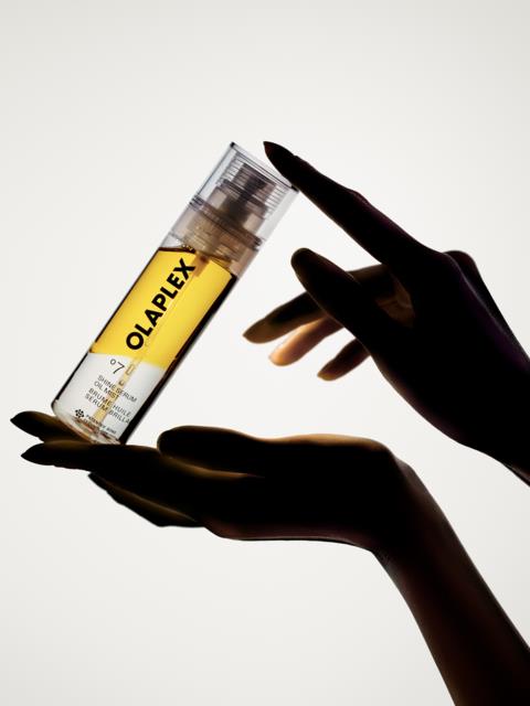 En hand som håller en genomskinlig flaska med gul vätska och pump, märkt Olaplex, mot en vit bakgrund.