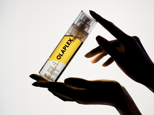 Hand som håller i en tydlig flaska med gul etikett som säger OLAPLEX, mot en ljus bakgrund.