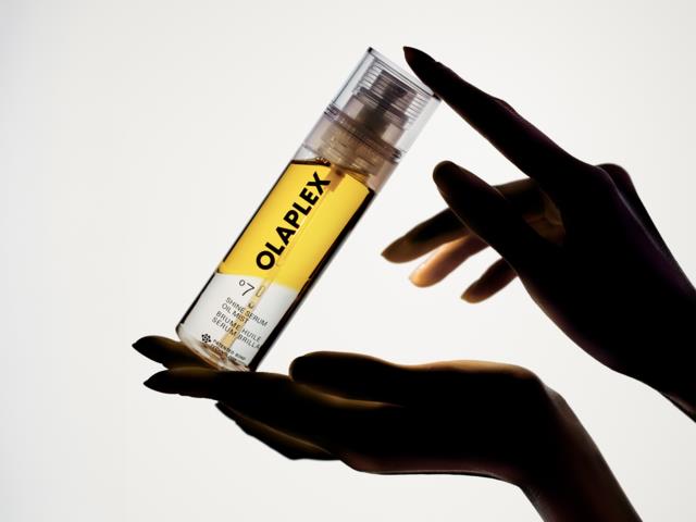 Hand som håller i en tydlig flaska med gul etikett som säger OLAPLEX, mot en ljus bakgrund.