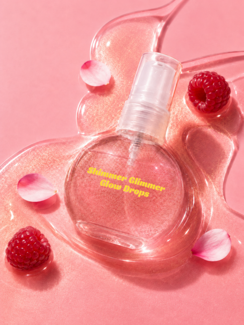 En genomskinlig sprayflaska märkt Shimmer Gummer Glow Drops omgiven av hallon och rosa blomblad på rosa bakgrund.