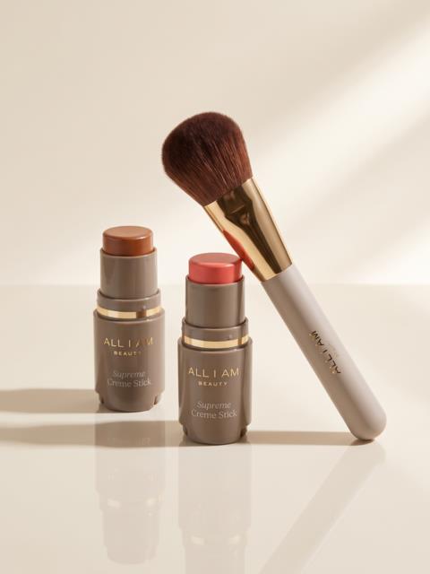 Två All I Am Beauty Supreme Creme Sticks i ljusa nyanser och en vit makeupborste med guldfattning.