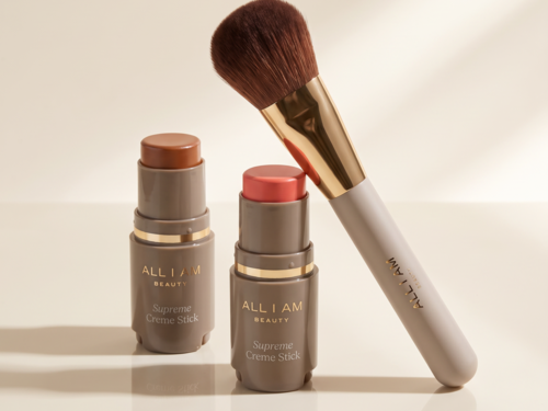 Två All I Am Beauty Supreme Creme Sticks i beige och rosa, och en makeupborste med vit skaft och guldfärgad detalj.