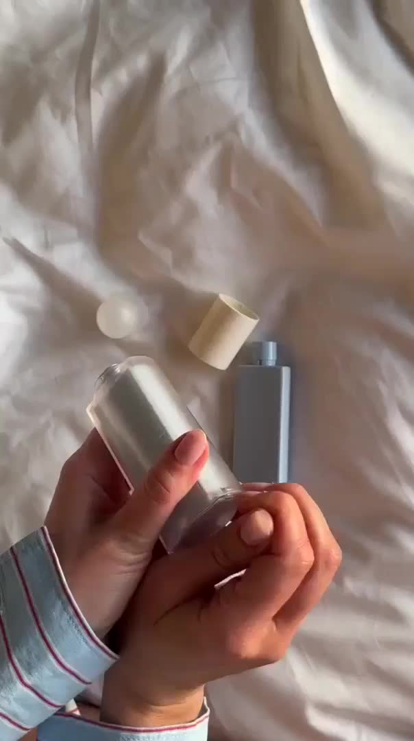 Två händer håller en silver sprayflaska och en liten vit behållare på vit tygbakgrund.