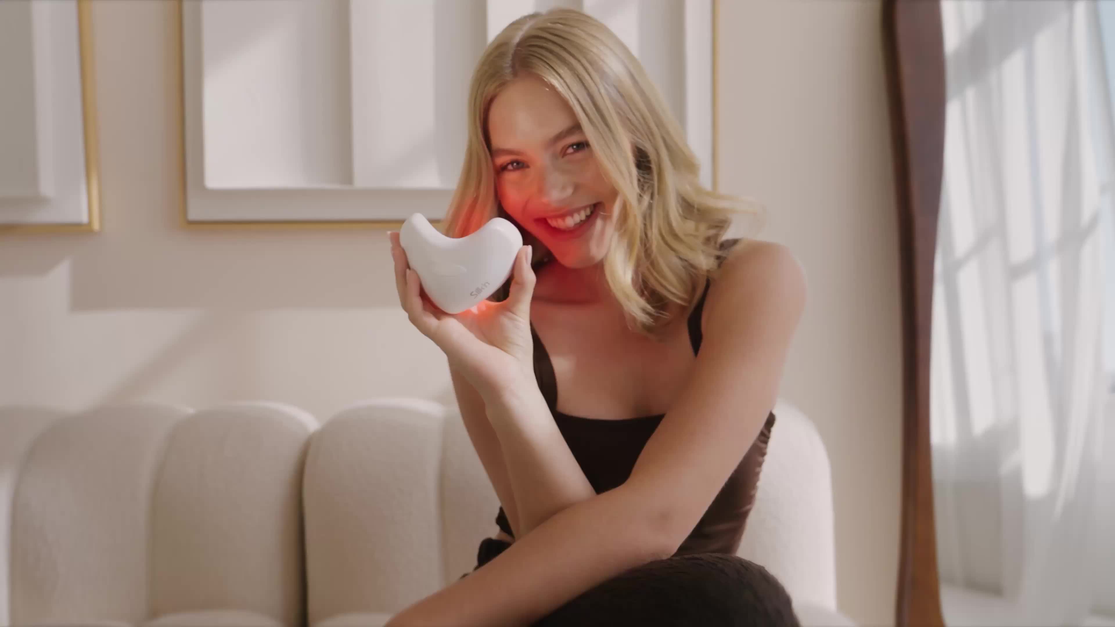 A woman smiling and holding a white object that looks like a small pillow or plushie in a cozy room.