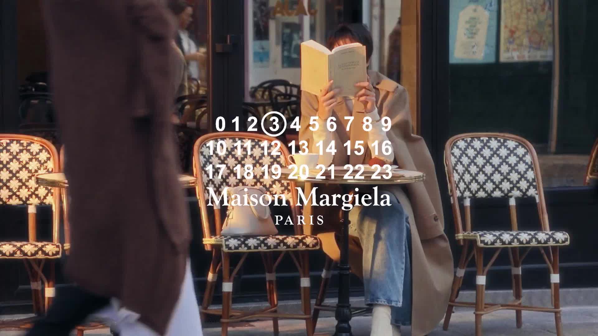Kvinna läser en bok på en gata, med stolar och en logotyp för Maison Margiela Paris i bakgrunden.