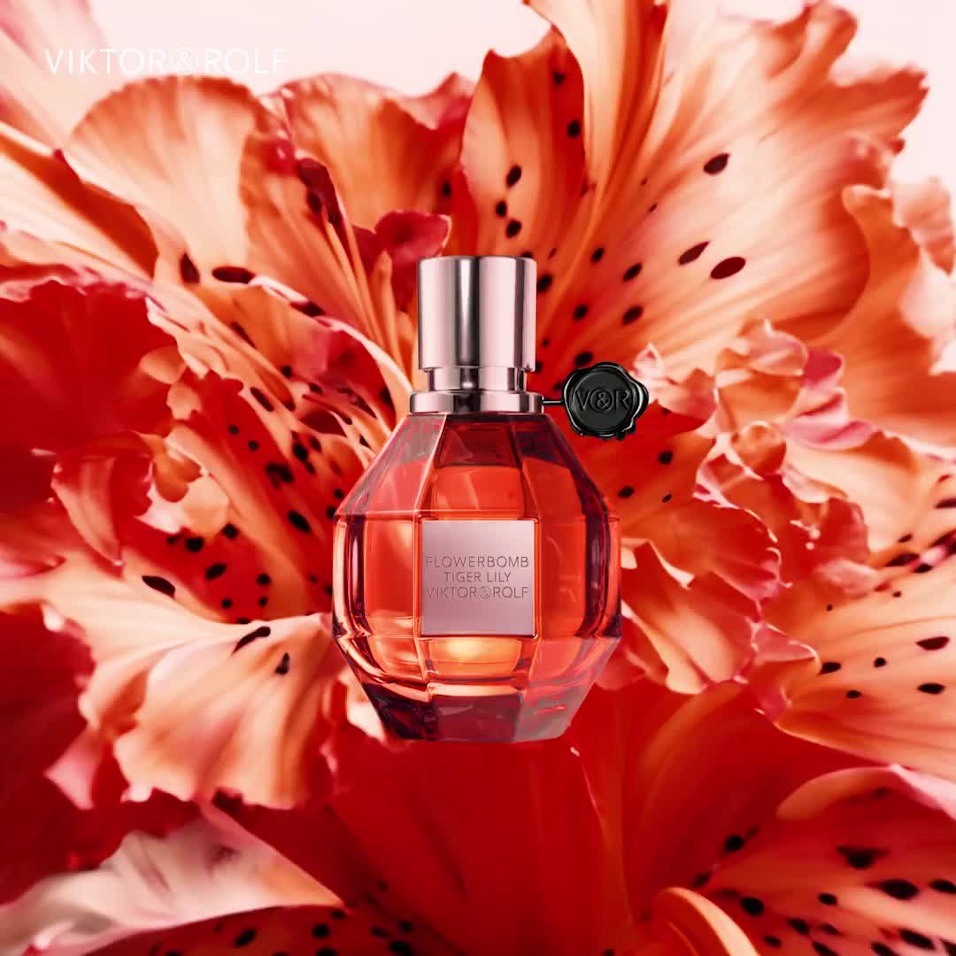 En parfymflaska med rosa vätska står framför en stor, livfull orange blomma. Flaskan har ett silverlock och en svart tag.